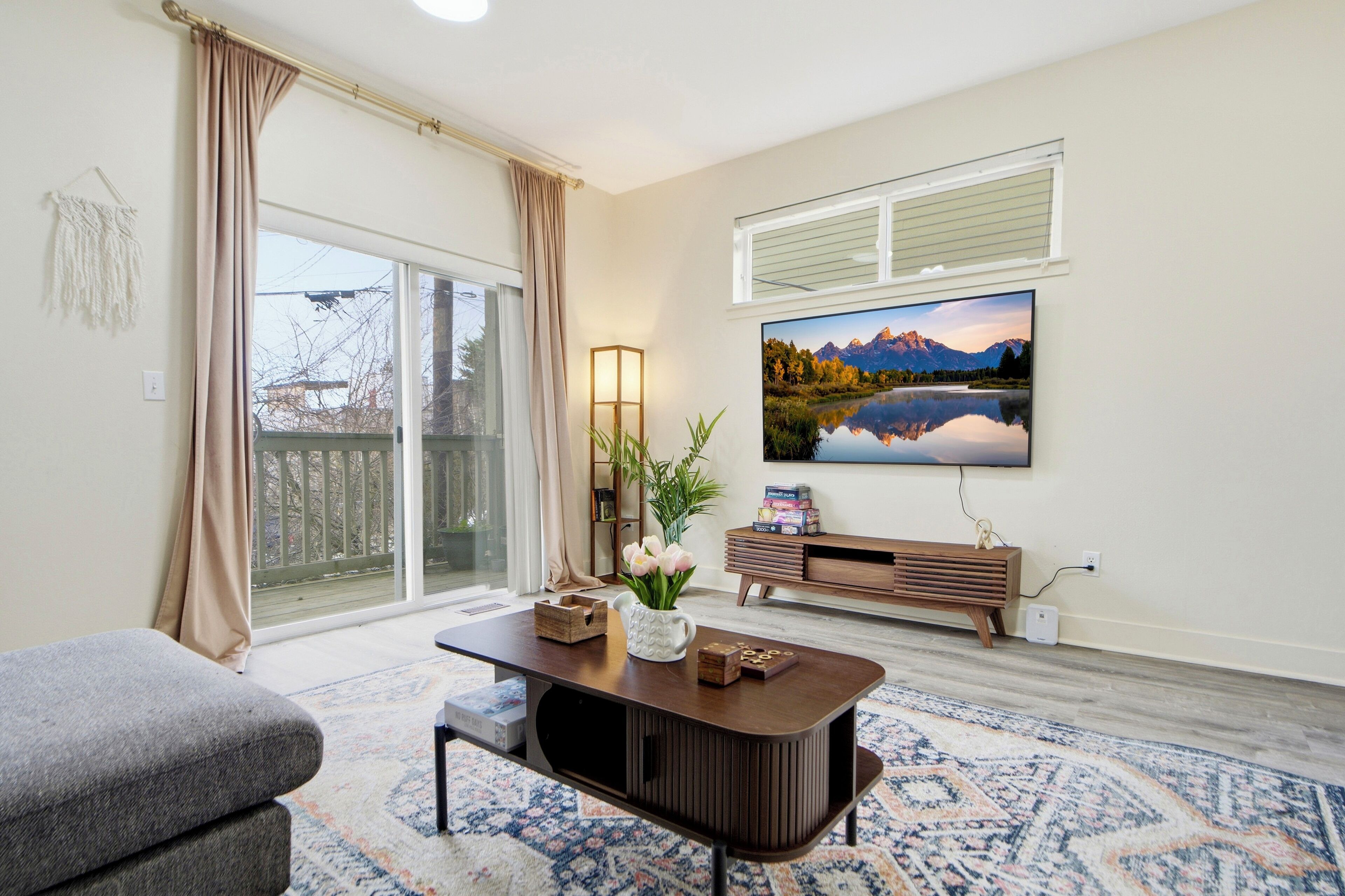 A living room with a large TV, a coffee table, and a sofa. There is a window with curtains, a plant, and a bookshelf.