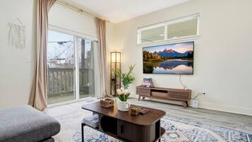 A living room with a large TV, a coffee table, and a sofa. There is a window with curtains, a plant, and a bookshelf.