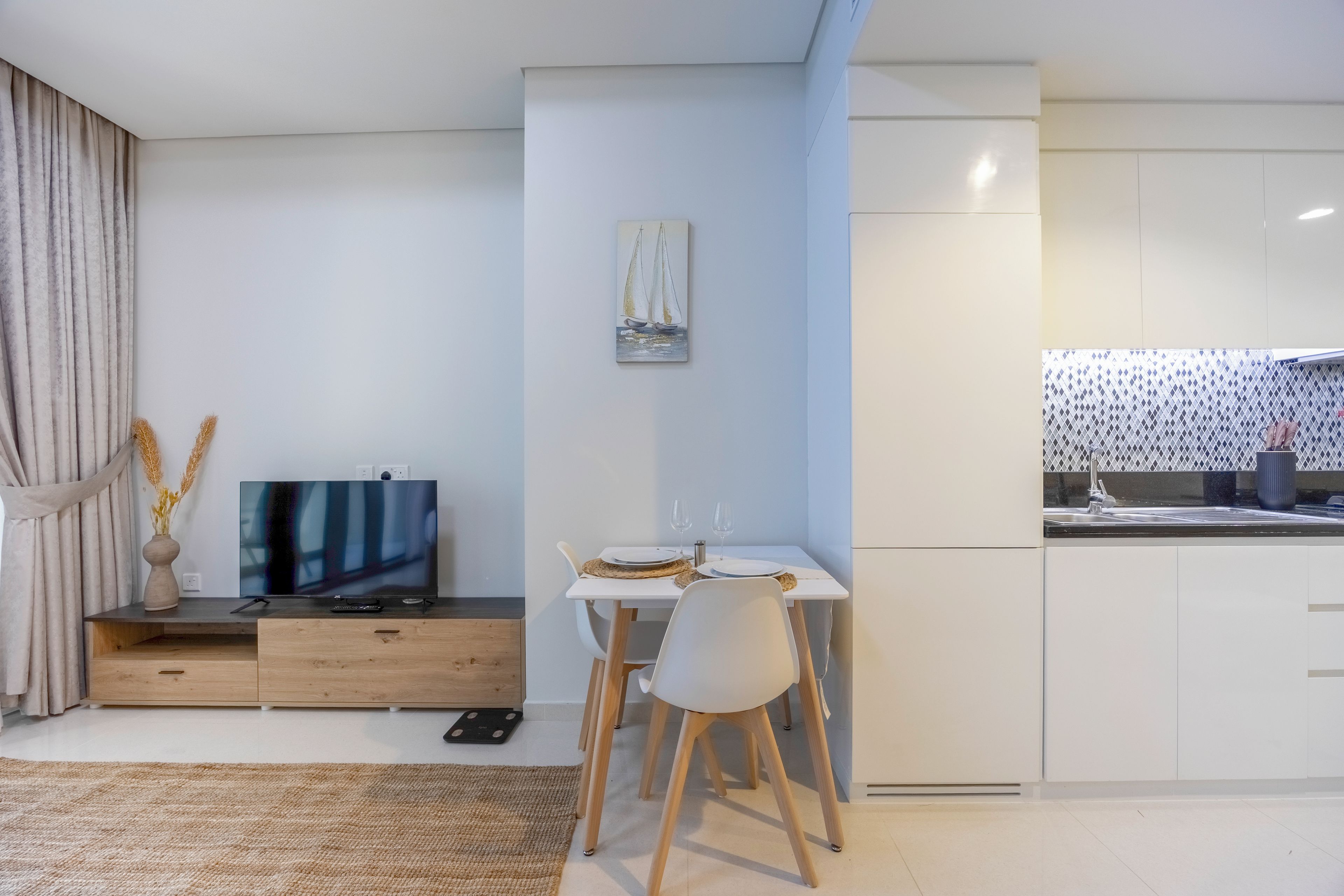 A modern living room with a flat-screen TV, a dining table with chairs, and a kitchen area with a sink and cabinets.