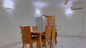 Ein Essbereich mit einem Glastisch, sechs Holzstühlen mit gemusterten Sitzbezügen, einem Kühlschrank und einem Deckenventilator.