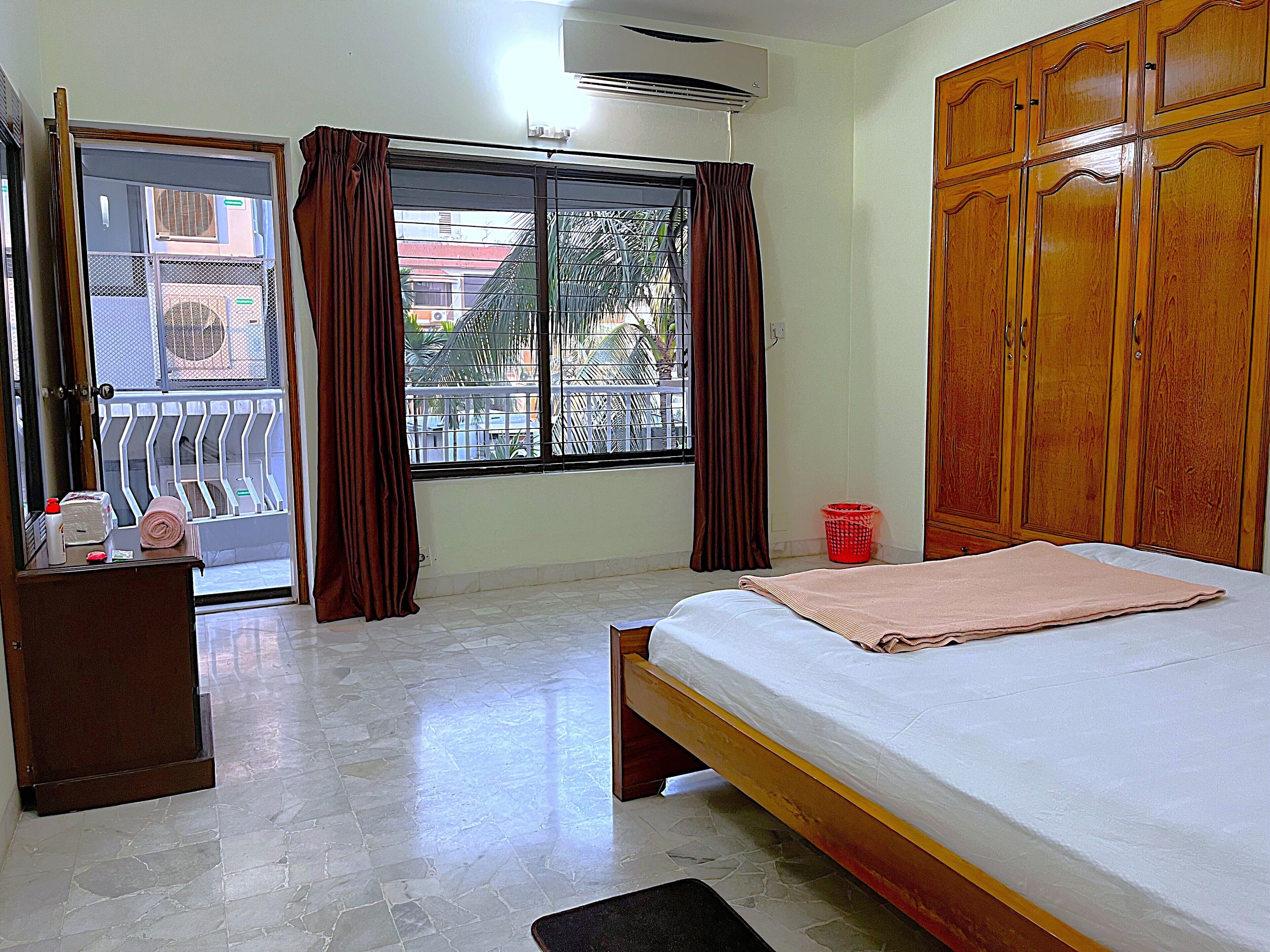 A bedroom with a bed, a wooden wardrobe, a desk with a chair, a window with curtains, and an air conditioning unit.
