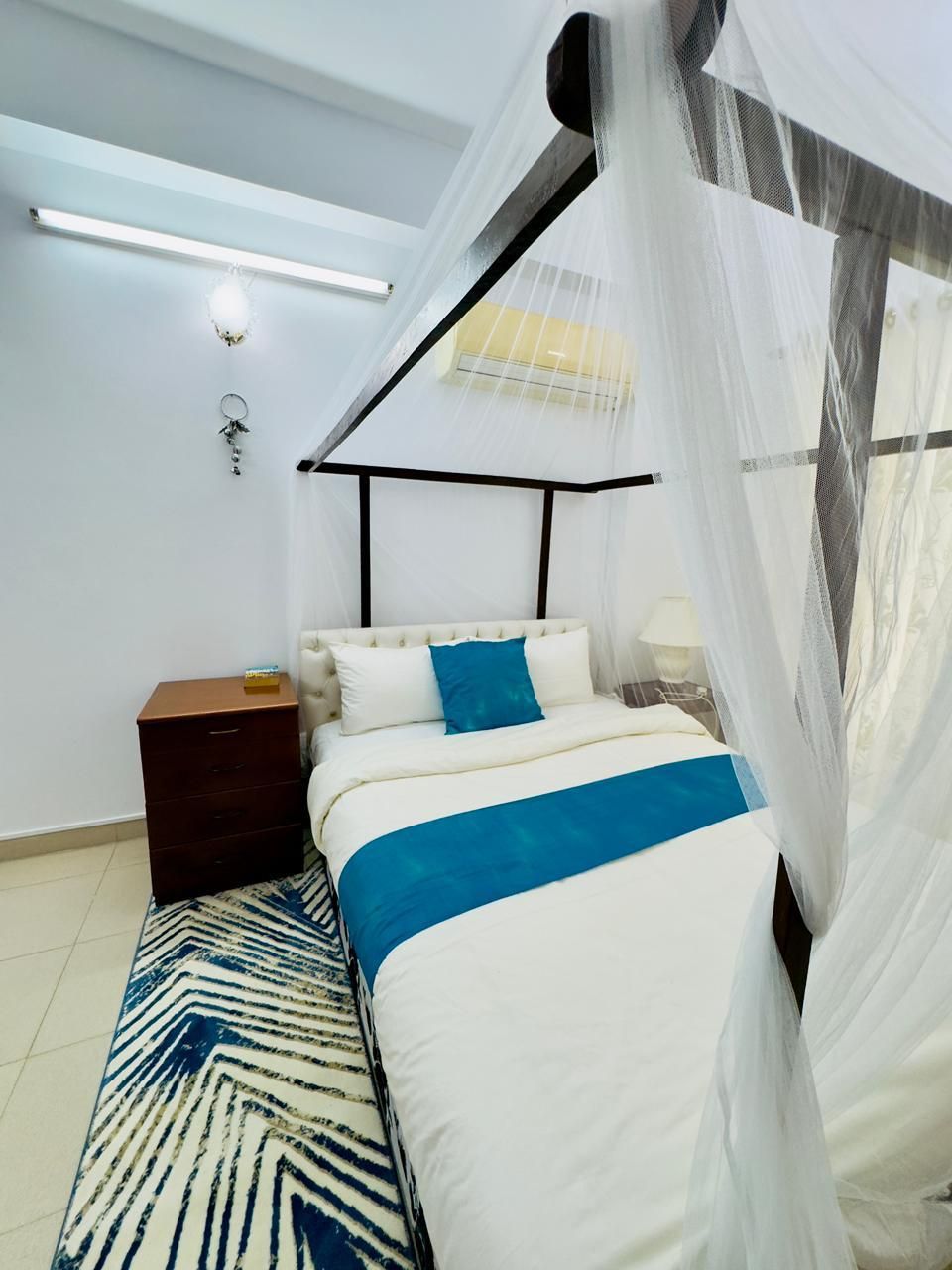 A bedroom with a bed, a nightstand, a bedside table, a lamp, and a window with a sheer curtain.