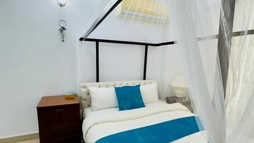 A bedroom with a bed, a nightstand, a bedside table, a lamp, and a window with a sheer curtain.