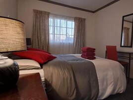 Una habitación de hotel con una cama, un escritorio, una lámpara, una silla y un ventanal con cortinas.