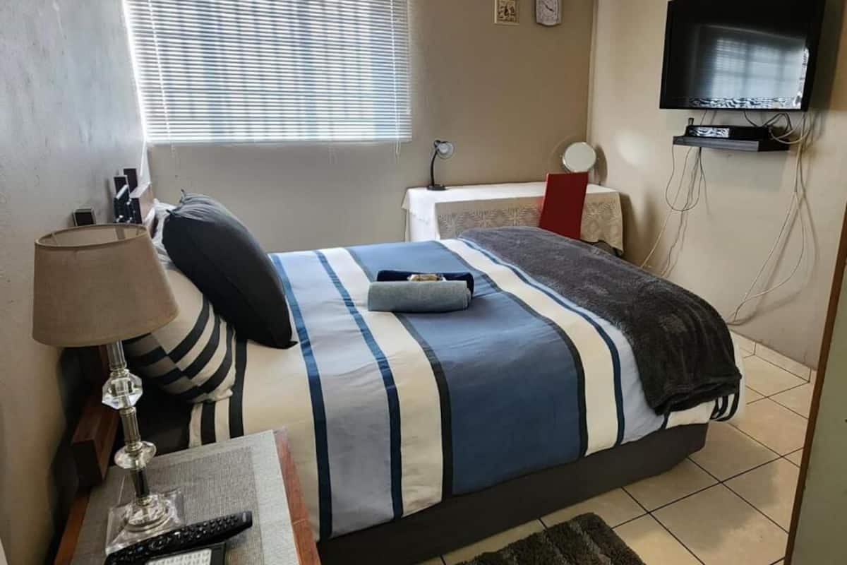 A bedroom with a bed, a TV, a bedside table, a lamp, and a window with blinds.