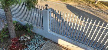 Un área cercada con valla metálica, un árbol y un pequeño jardín con flores rojas y follaje azul.