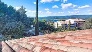 Eine Küstenstadt mit terrakottafarbenen Dächern, einem markanten, hohen Baum und Gebäuden mit Balkonen mit Blick aufs Meer.