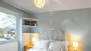 Un dormitorio con una cama, dos mesitas de noche con lámparas, un ventanal con vistas a casas y una estantería con fotografías.