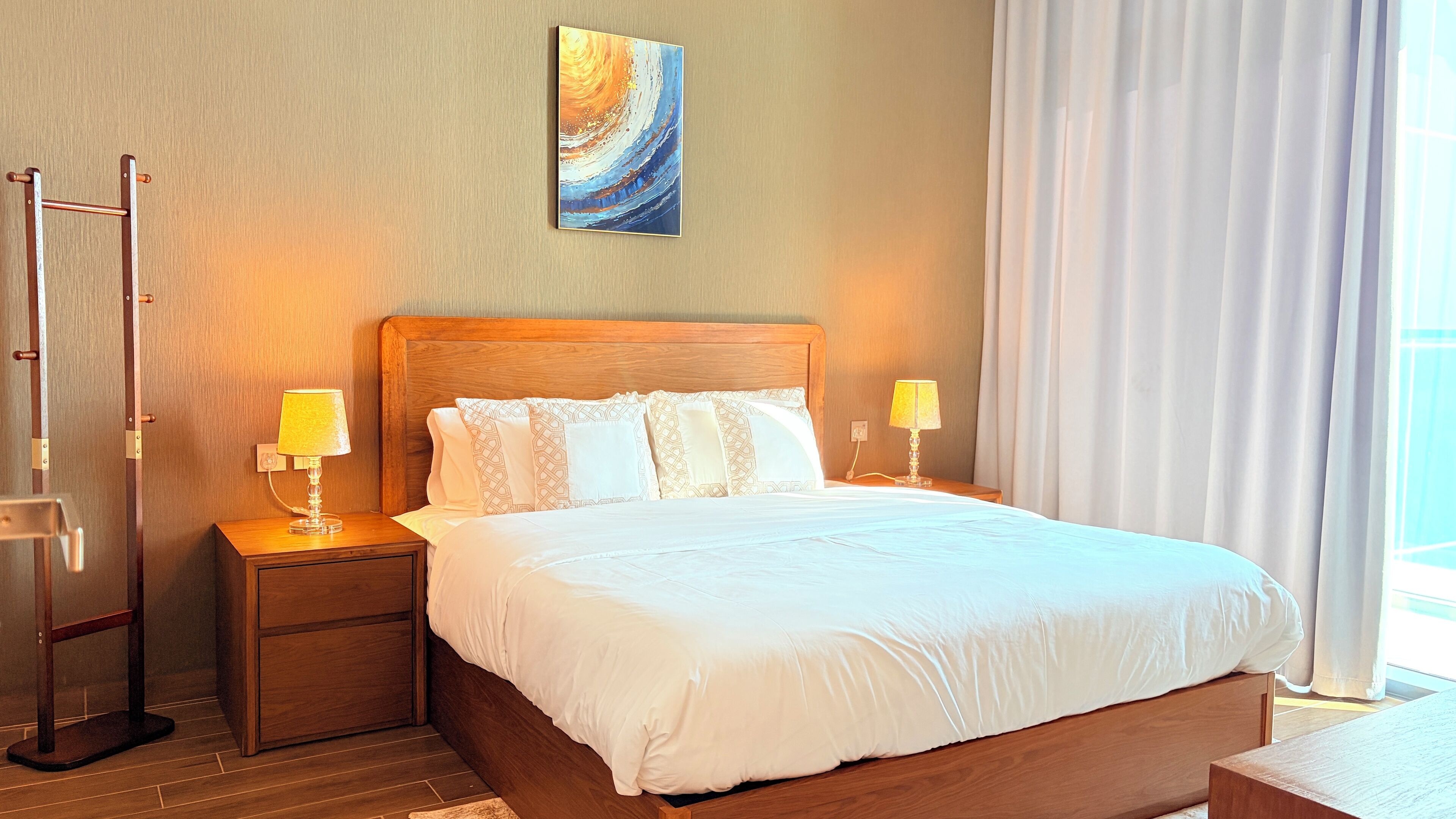 Une chambre d’hôtel avec un lit en bois, deux lampes de chevet, une table de chevet en bois, un plancher en bois, un tableau au mur et une fenêtre avec des rideaux.