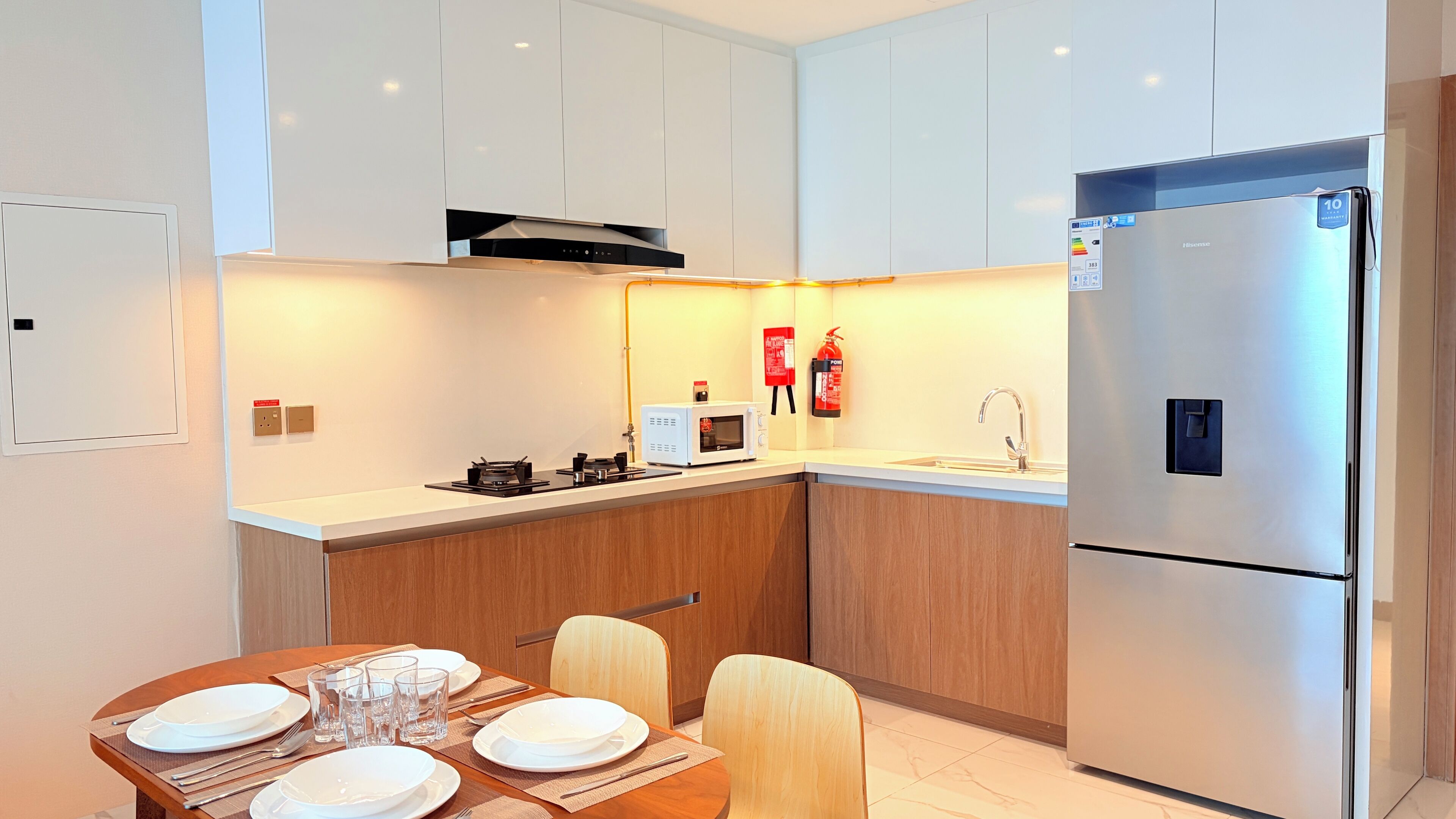 Een moderne keuken met witte kasten, een roestvrijstalen koelkast, een magnetron en een gasfornuis. Er staat een eettafel gedekt met borden en glazen.