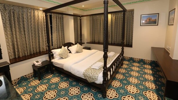 Una cama con dosel de madera, ropa de cama blanca y almohadas estampadas. La habitación tiene alfombra estampada y cortinas con borde verde.