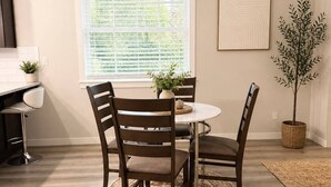 Un coin repas avec une table ronde, quatre chaises, un lustre, une fenêtre avec des stores, une plante en pot et un tapis.