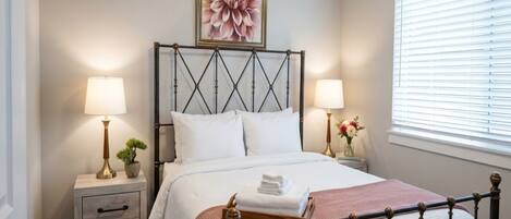A neatly made bed with a headboard, two bedside tables with lamps, a wall-mounted flower painting, and a window with blinds.