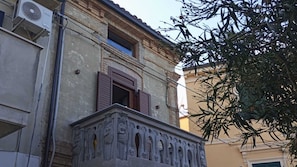 Un edificio con un balcón que presenta elaboradas esculturas, una ventana con contraventanas de madera y una unidad de aire acondicionado instalada en la pared.
