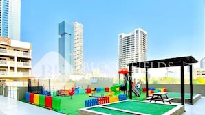 An outdoor playground with slides and climbing structures, surrounded by a paved area and buildings in the background. - Bloomfields Modern Escape Stu In Jvc (Dubai)