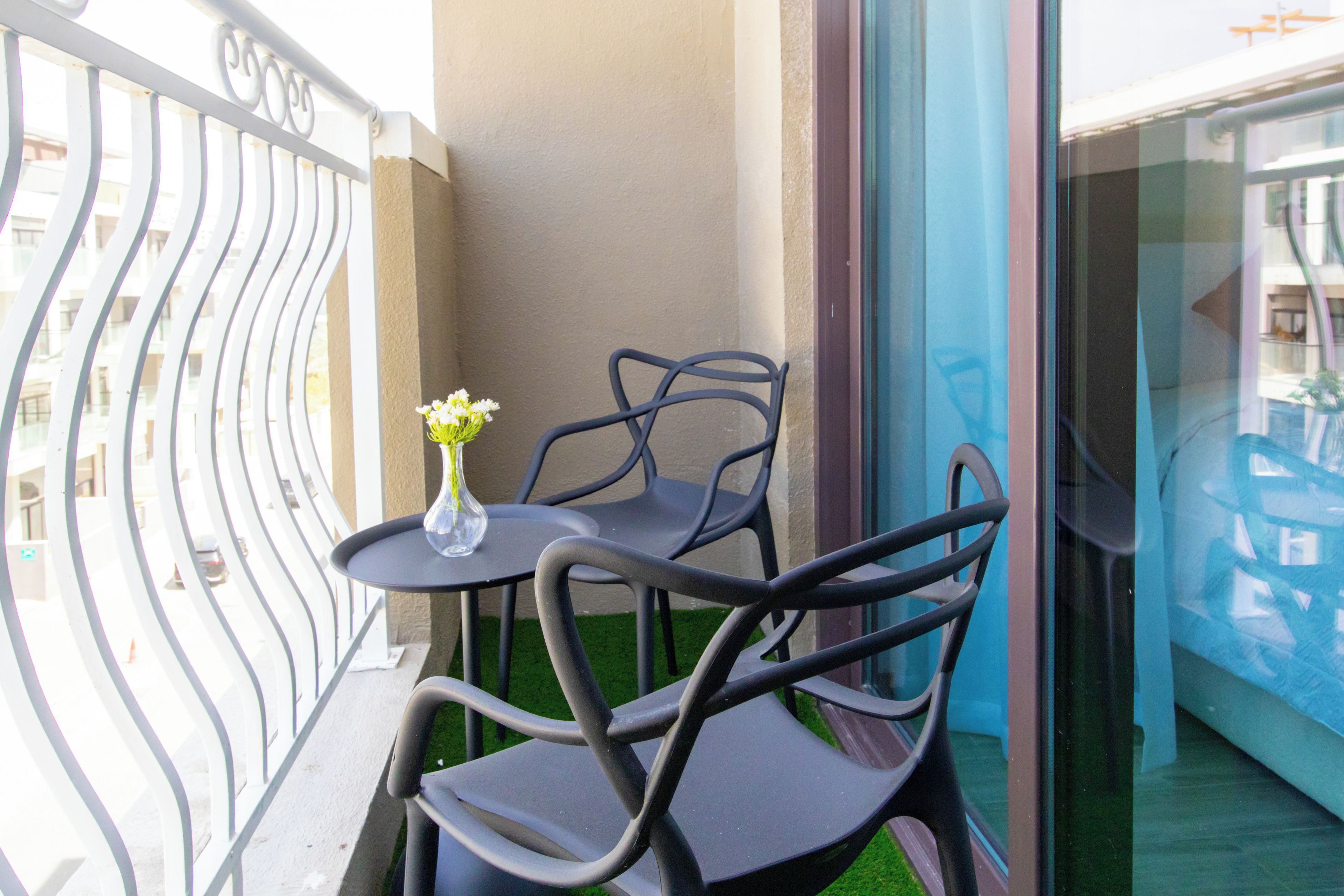 Varanda com mesa e duas cadeiras, um vaso de flores e vista de um edifício através de uma porta de vidro.