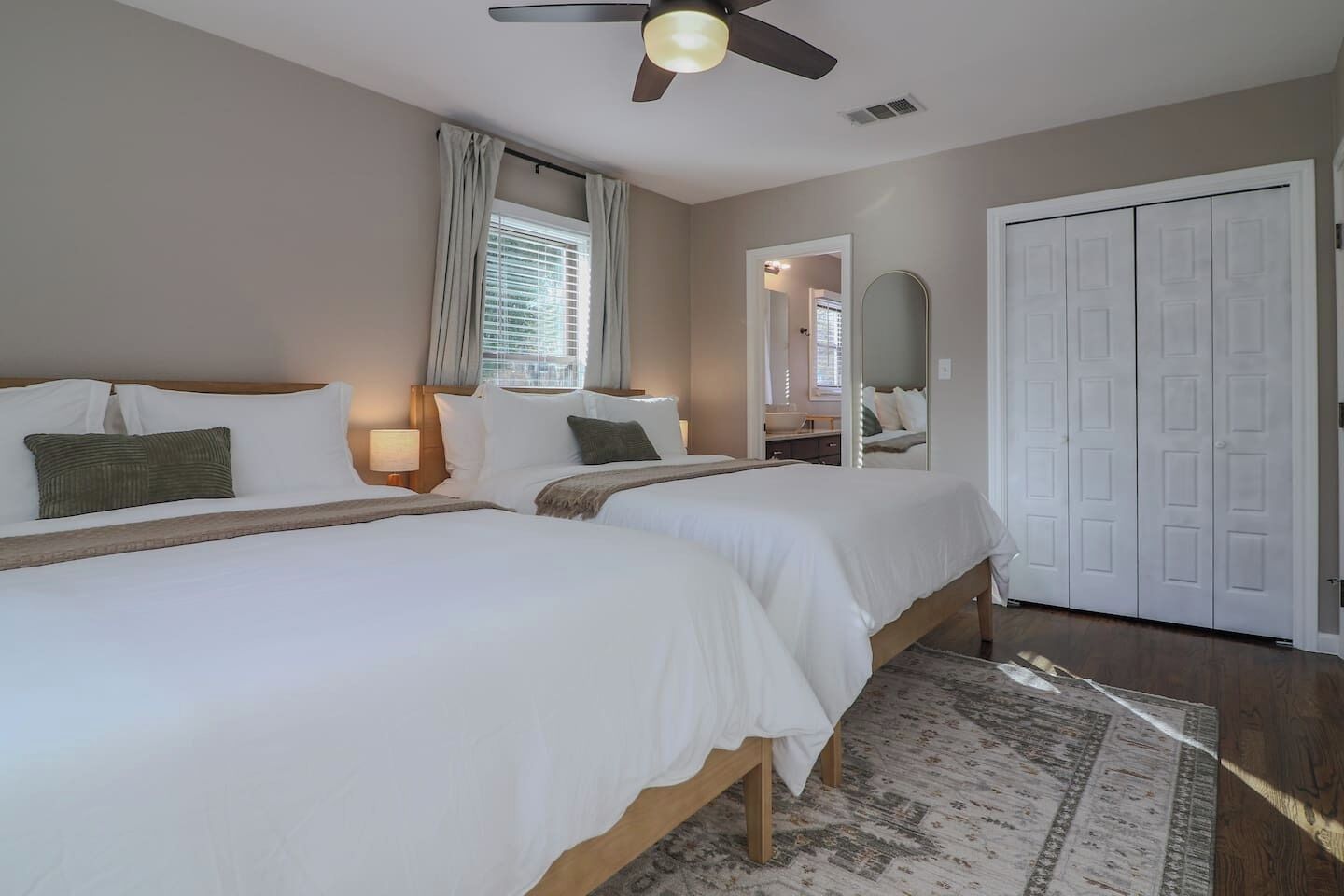 A hotel room with two beds, a ceiling fan, a window with curtains, a mirror, and a wardrobe.