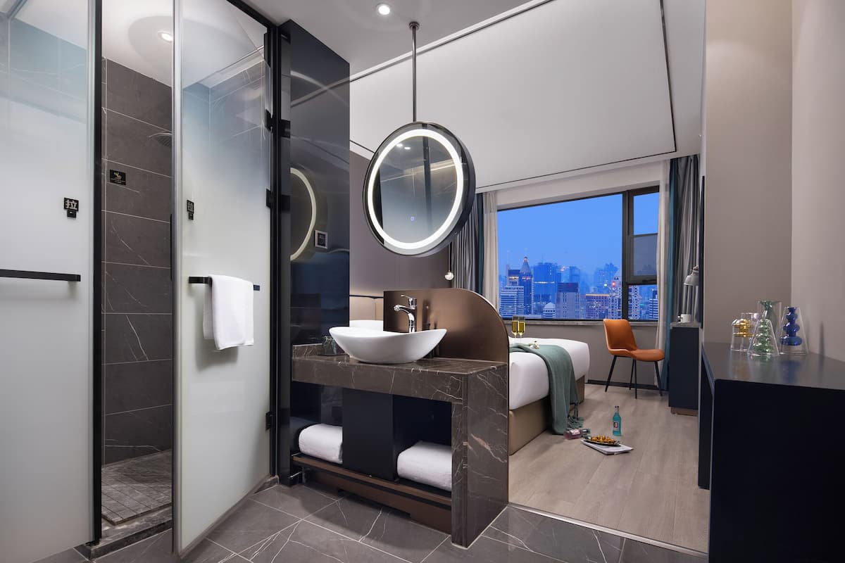 Une salle de bain d’hôtel moderne avec une douche à parois de verre, un meuble-lavabo en marbre doté d’un grand évier, un miroir rond et une vue sur la ville par la fenêtre.
