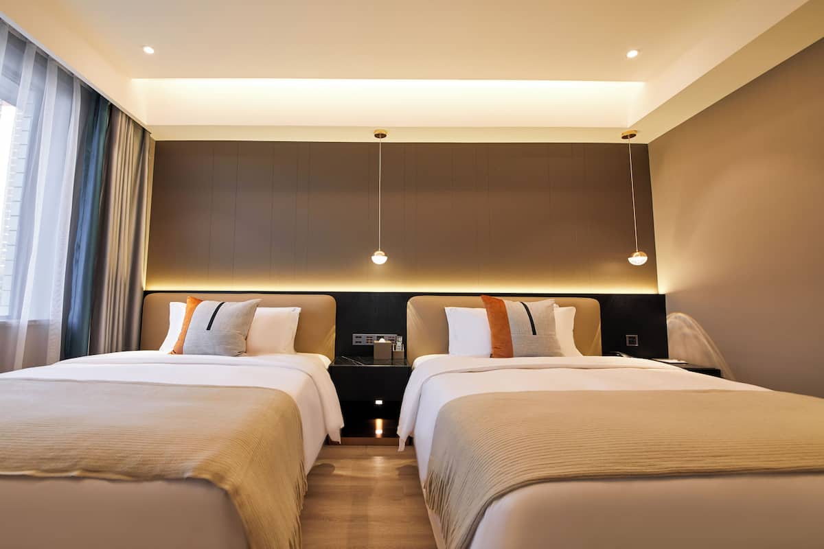 Une chambre d’hôtel avec deux lits, une table de chevet, une lampe et une vue sur la ville à travers des rideaux transparents.