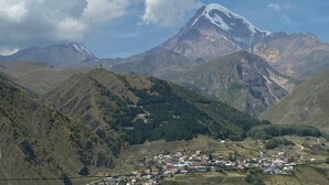 Une montagne dont le sommet est coiffé de neige, une vallée abritant un petit village, et un ciel dégagé parsemé de nuages.