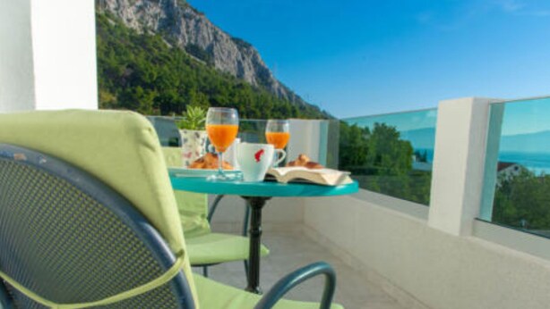 A balcony with a table set for breakfast, including orange juice and a book, overlooking a mountainous landscape.