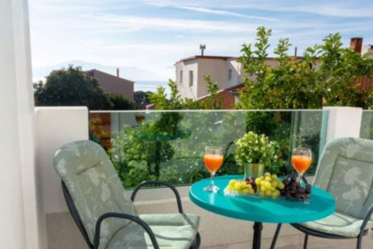 A balcony with a table set for two, featuring two glasses of orange juice, a fruit basket, and a vase with flowers.