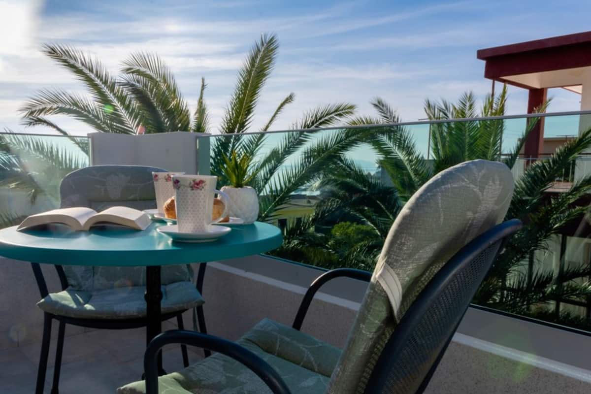 A round table with chairs, a book, and a floral-patterned cup set on a balcony with palm trees in the background.
