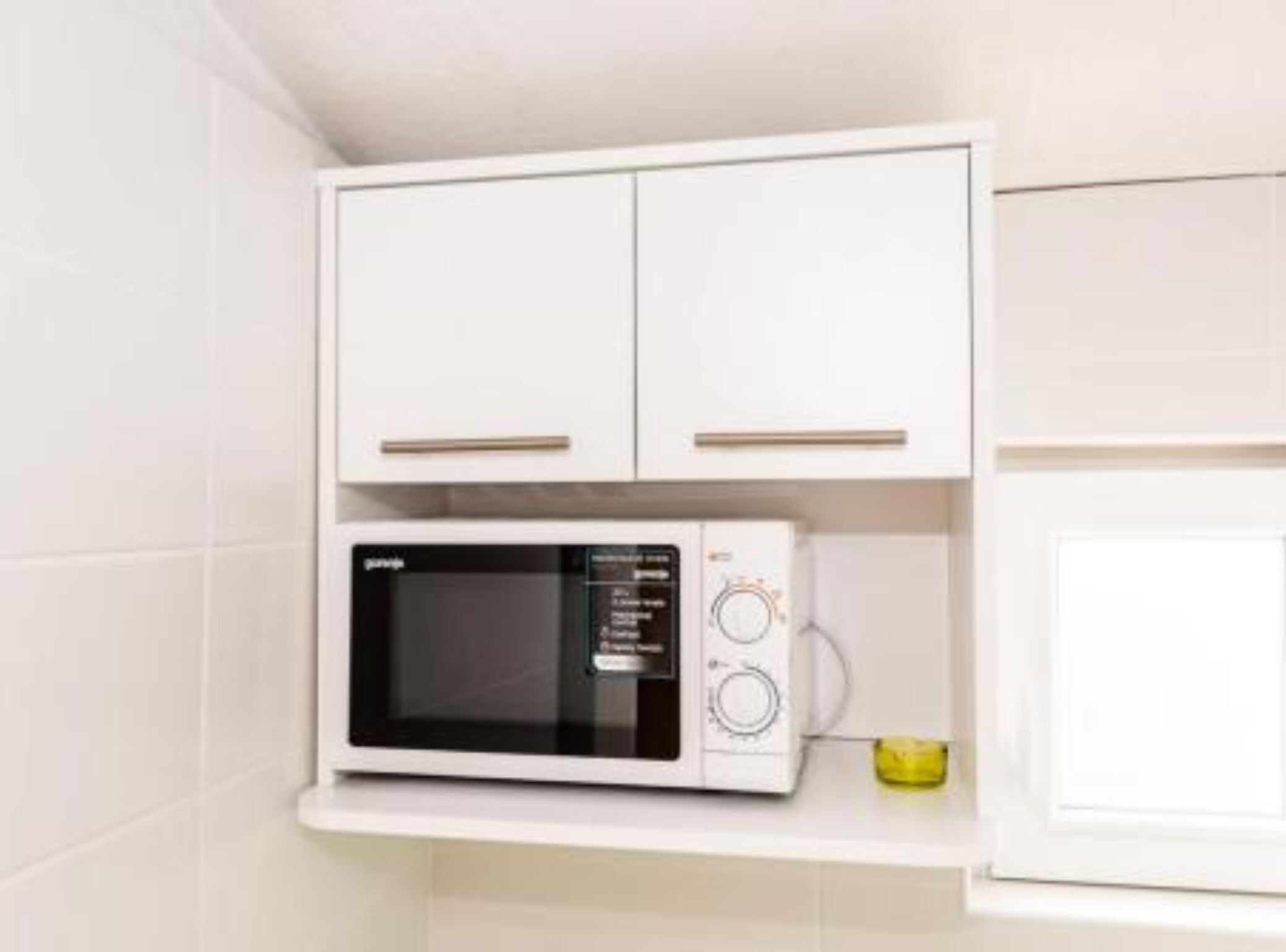 Un microondas blanco sobre una repisa blanca en una cocina con armarios blancos.