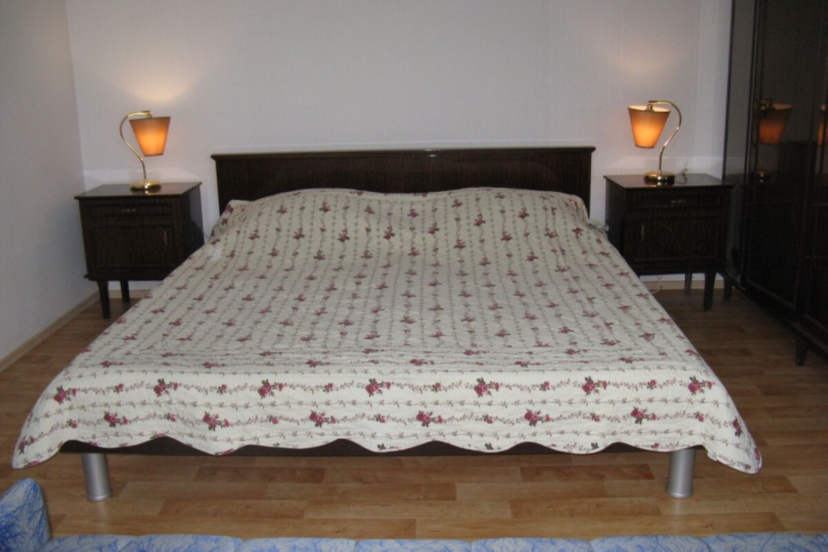 A bed with a floral-patterned bedspread, two bedside tables with lamps, and a wooden floor.