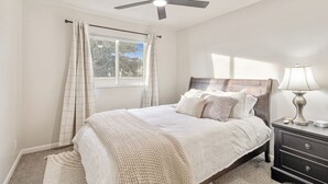 Une chambre à coucher avec un lit, une table de chevet, une lampe, un ventilateur de plafond et une fenêtre avec des rideaux.