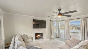 Un salon doté d’une cheminée, d’un ventilateur de plafond et d’une grande fenêtre avec des rideaux.