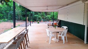 Een terras buiten met een overdekte zithoek, een ronde tafel met stoelen en uitzicht op een tuin.