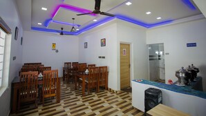 A dining area with wooden chairs and tables, a ceiling fan, and a kitchen counter with appliances. - Hazeberg Resort Munnar (Devikolam)