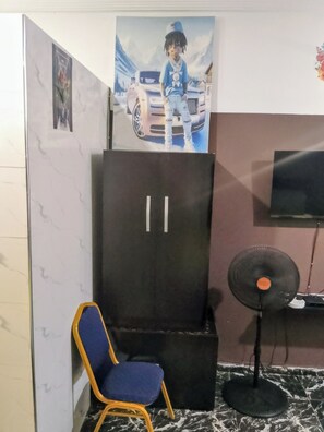 Ein Zimmer mit einem dunkelbraunen Schrank, einem blauen Stuhl, einem schwarzen Ventilator und einem Fernseher an der Wand.