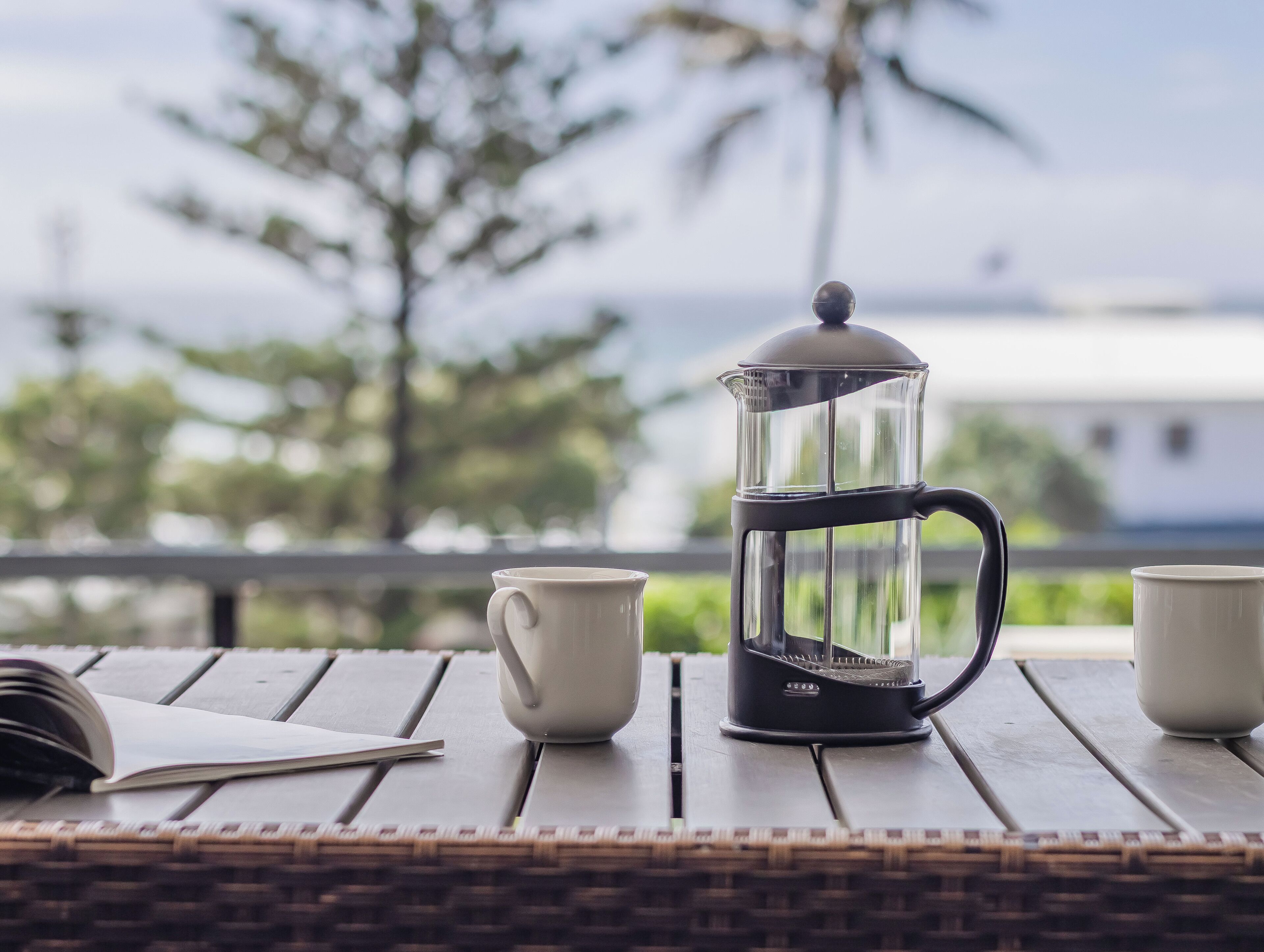 A coffee maker on a wooden table with a book and two cups, set outdoors with a view of trees and a building.