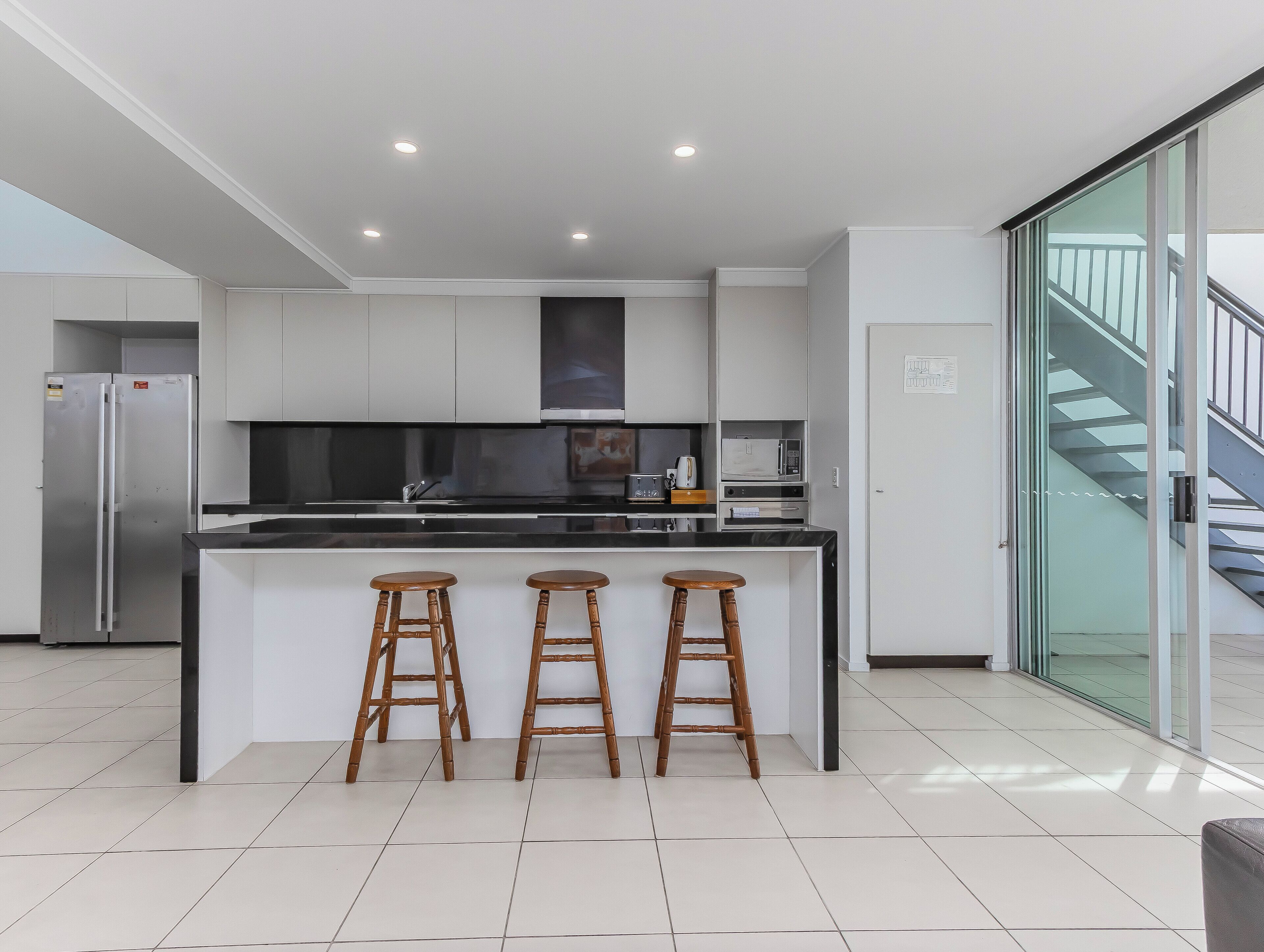 A modern kitchen with a central island, bar stools, stainless steel appliances, and a glass door leading to a staircase.