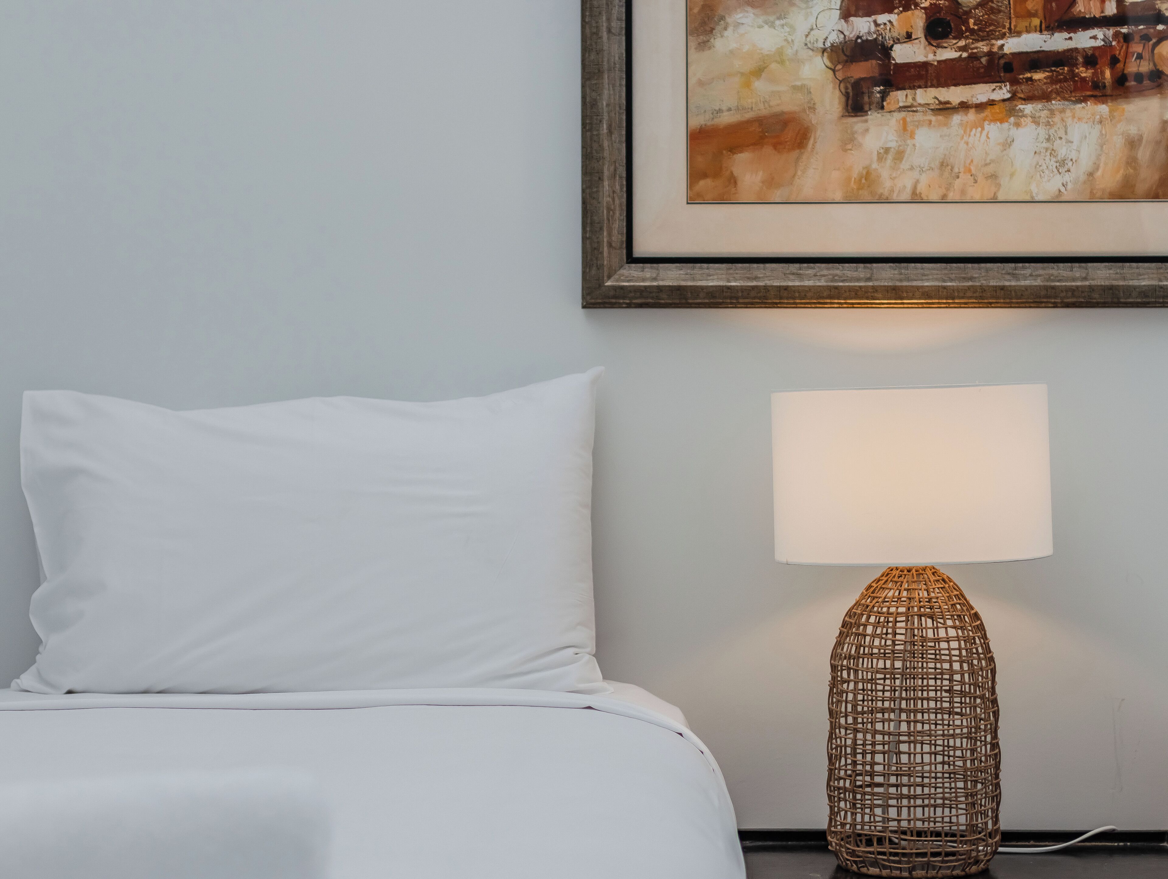 A bed with white bedding, a single white pillow, a wall-mounted lamp, and a framed abstract painting.