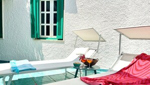 Et solbadet rum med en pool, to liggestole, et bord med en drik og et vindue med grønne skodder.