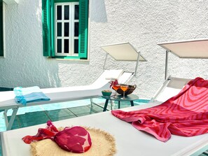 Ein sonnendurchfluteter Raum mit Pool, zwei Liegestühlen, einem Tisch mit einem Getränk und einem Fenster mit grünen Rollläden.