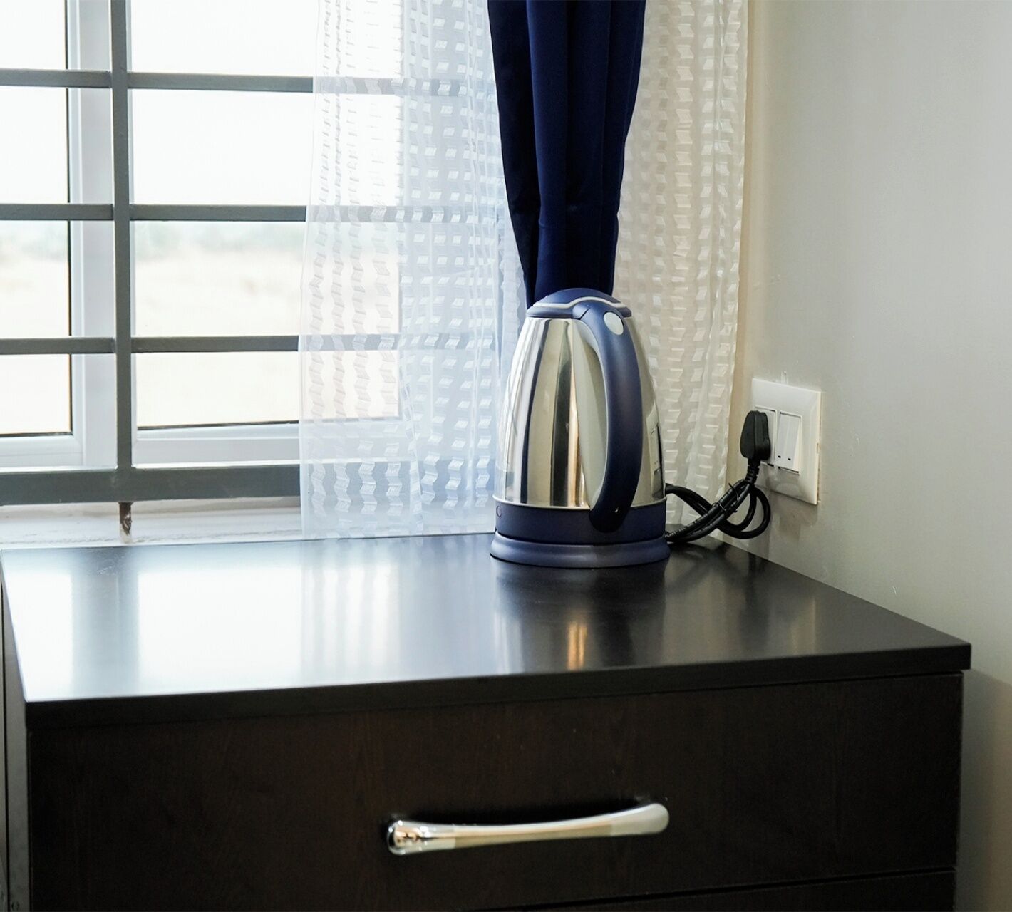 A kettle on a dark wooden table near a window with a white power outlet.