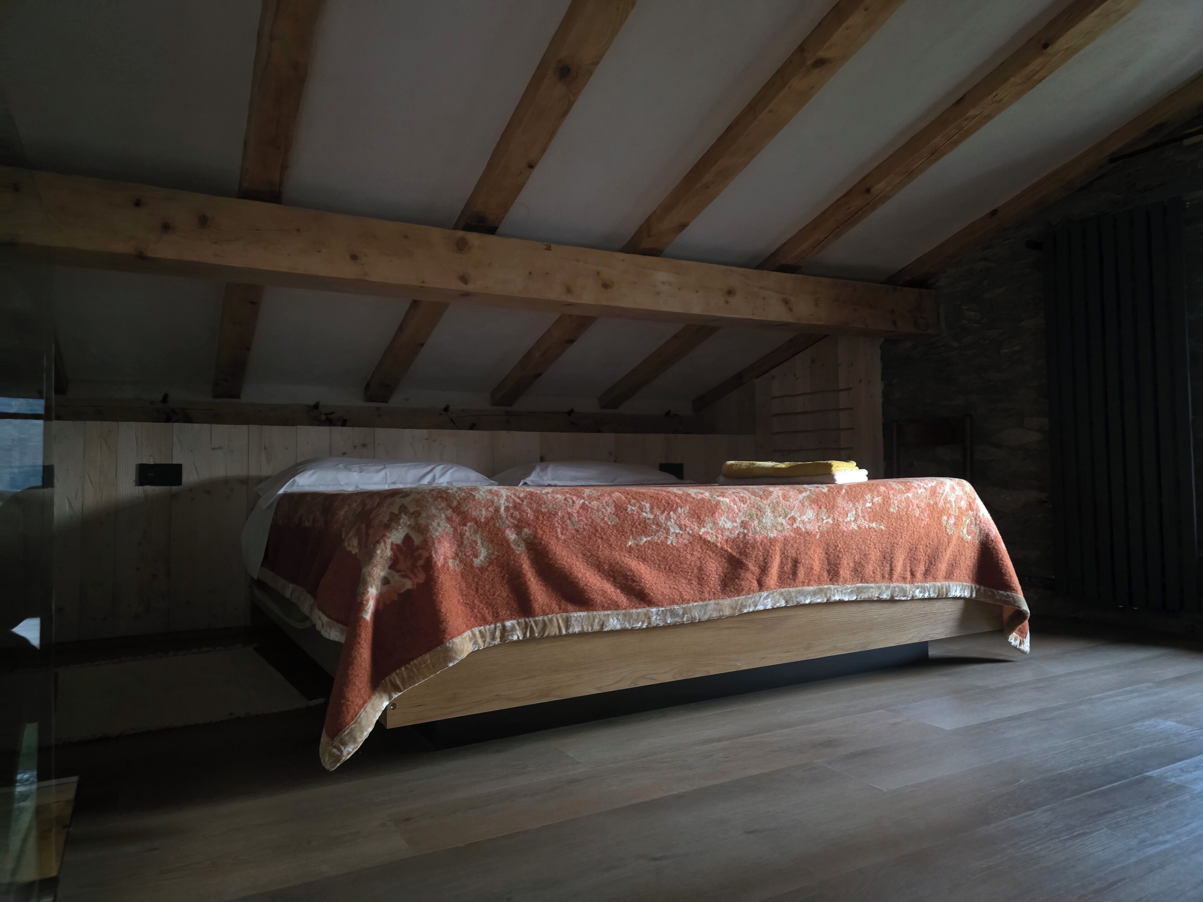 A bedroom with a wooden ceiling and sloped roof, a bed with a red blanket, and a stone wall.