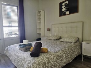 Ein ordentlich bezogenes Bett mit gemusterten Textilien, ein Nachttisch, eine Lampe und ein an der Wand hängendes Kunstwerk.