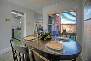 A dining area with a round table set for four, a kitchen in the background, and a window with blinds. - Coastal Condo + Near Oso Bay & Padre Beaches | Seaside Haven (Corpus Christi)