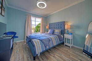 Ein Schlafzimmer mit Bett, Schreibtisch, Stuhl, einem Fenster mit Vorhängen und einem Nachttisch mit Lampe.