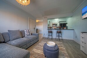 Un salon avec un canapé d’angle gris, un pouf rond et un coin cuisine comprenant un tabouret de bar et un four à micro-ondes.