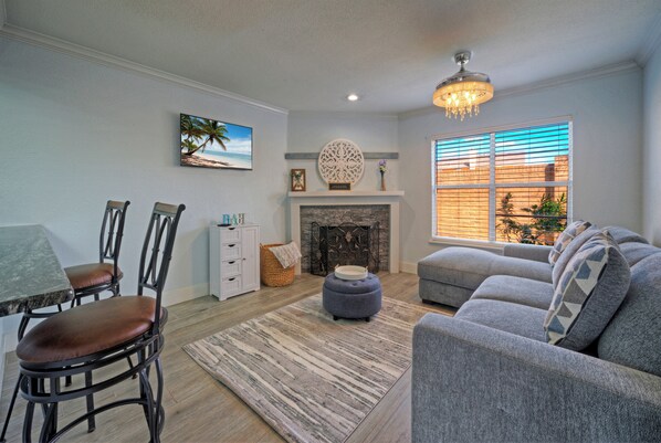 A living room with a fireplace, a dining table with chairs, a sofa, and a large window with blinds. - Coastal Condo + Near Oso Bay & Padre Beaches | Seaside Haven (Corpus Christi)