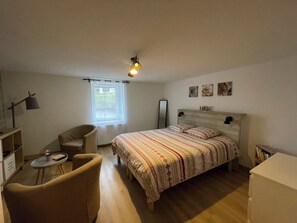 Un dormitorio con cama, una silla, una mesita, un espejo y una ventana.