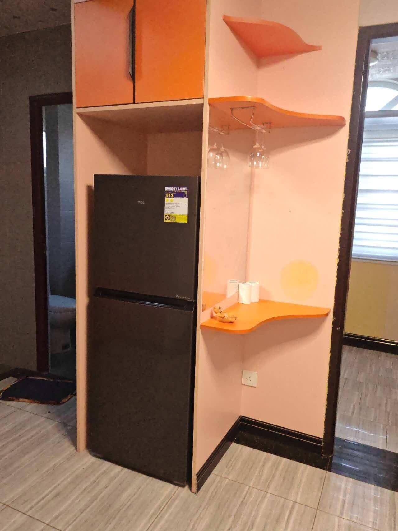 A compact kitchen with a black refrigerator, a countertop with a glass jar, and a wall-mounted shelf with a glass object.