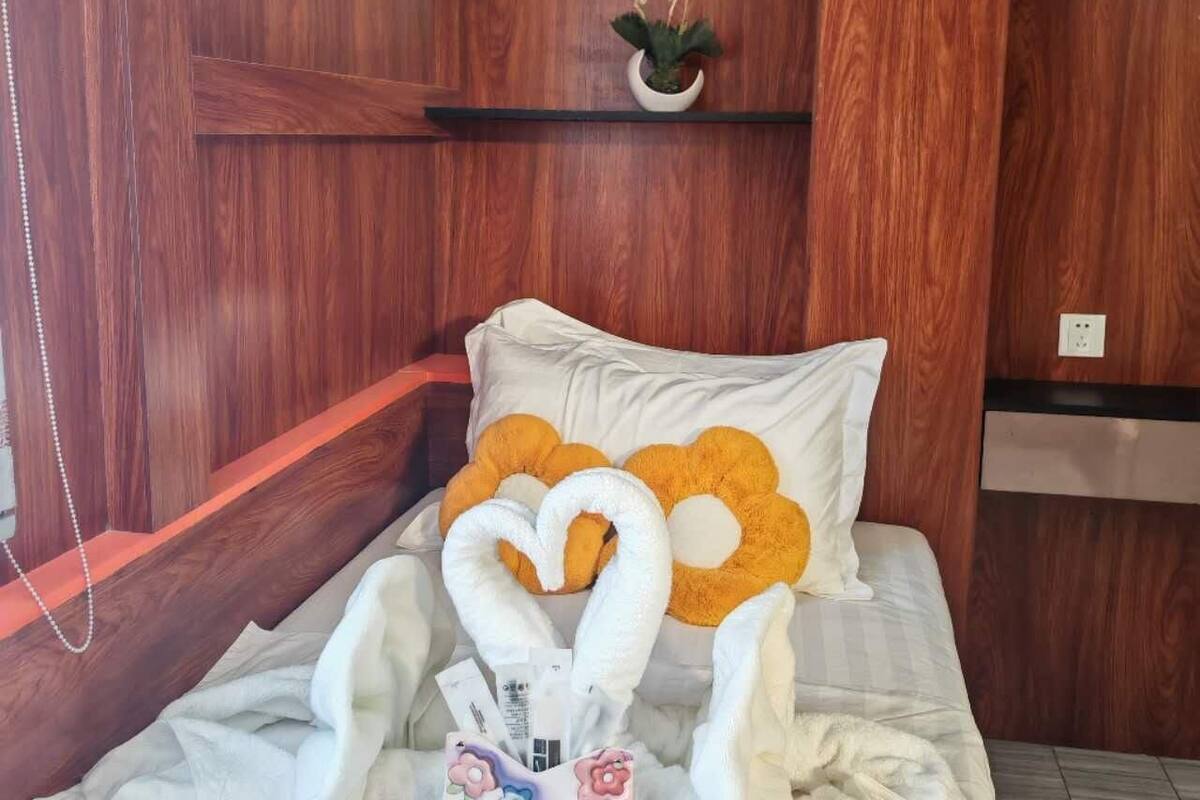 A neatly made bed with white bedding and decorative pillows, a small potted plant on a shelf, and a wall-mounted air conditioner.