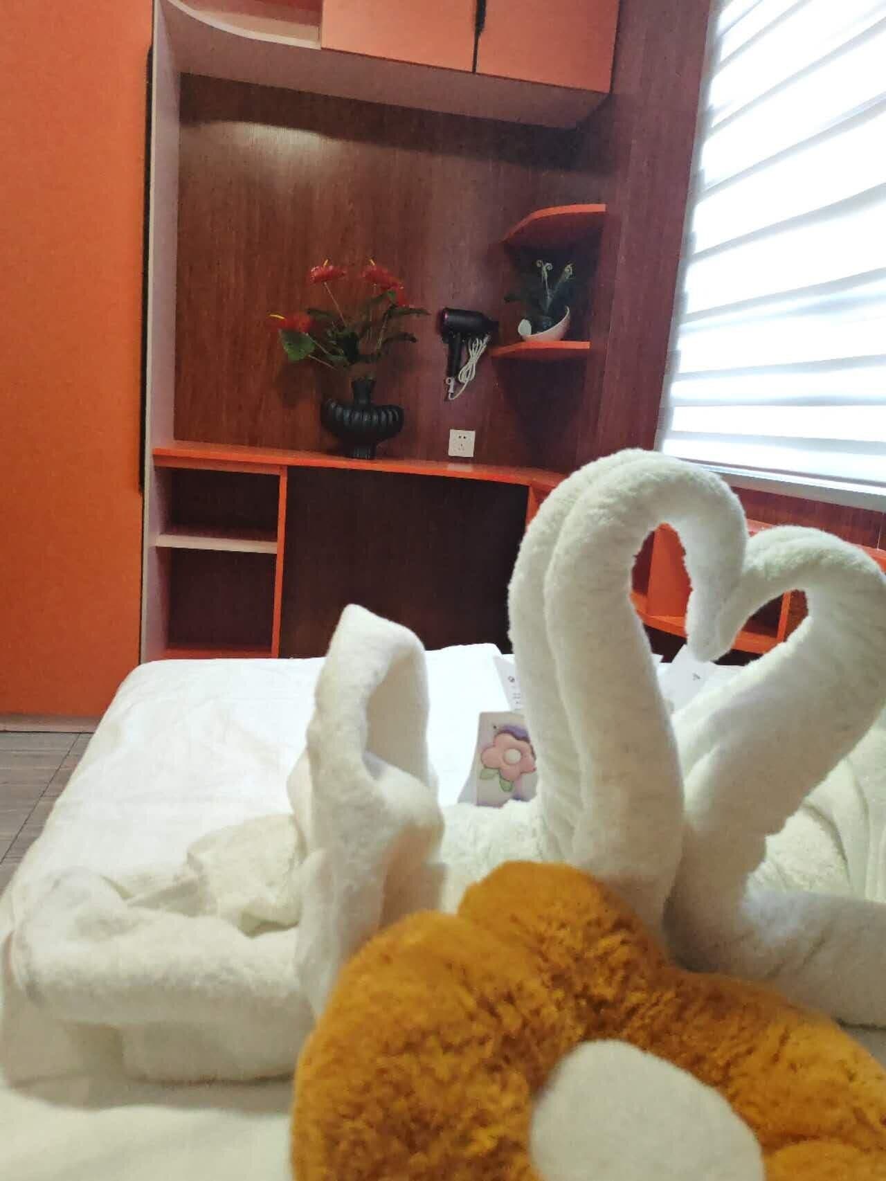 Un lit avec des serviettes blanches disposées en forme de cœur, un jouet en peluche et une étagère en bois sur laquelle se trouvent une plante en pot et un sèche-cheveux.